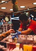 A fruit smoothie vendor is handing a drink to a customer at a market stall. The vendor is wearing a red shirt and blue gloves, while the customer is reaching out for the drink. Various colorful smoothies and icy drinks are displayed on the counter, with a hanging pineapple above and signs showing the different flavors.