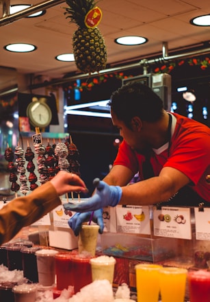A fruit smoothie vendor is handing a drink to a customer at a market stall. The vendor is wearing a red shirt and blue gloves, while the customer is reaching out for the drink. Various colorful smoothies and icy drinks are displayed on the counter, with a hanging pineapple above and signs showing the different flavors.