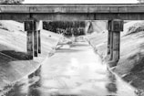 A black and white photograph of a concrete drainage canal with a bridge crossing over it. The canal is lined with smooth concrete walls, and graffiti is visible on the bridge's supporting columns. The bridge is constructed of concrete with visible weathering, and beyond the bridge, there are three large culverts.