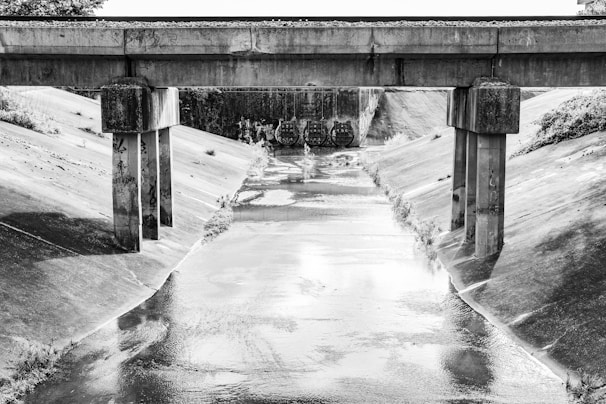 A black and white photograph of a concrete drainage canal with a bridge crossing over it. The canal is lined with smooth concrete walls, and graffiti is visible on the bridge's supporting columns. The bridge is constructed of concrete with visible weathering, and beyond the bridge, there are three large culverts.