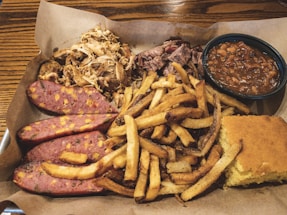 A hearty meal featuring sliced sausage with visible cheese chunks, seasoned fries, shredded meat, baked beans, and a piece of cornbread presented on a rustic brown paper.