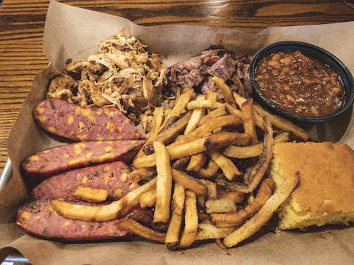 A hearty meal featuring sliced sausage with visible cheese chunks, seasoned fries, shredded meat, baked beans, and a piece of cornbread presented on a rustic brown paper.