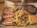 A hearty meal featuring sliced sausage with visible cheese chunks, seasoned fries, shredded meat, baked beans, and a piece of cornbread presented on a rustic brown paper.