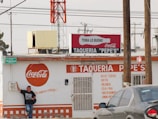 A street taco shop named 'Taqueria Pepe's' with a prominent Coca-Cola branding on its exterior. The building appears weathered and is painted primarily in white and orange. A person stands casually against the wall on the left, wearing jeans and a black t-shirt. There are several signs, including a green street sign reading 'Higuera'. Overhead, power lines are visible with a large red and white tower in the background. A gray car is parked in front on the right side.