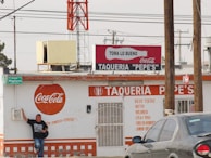 A street taco shop named 'Taqueria Pepe's' with a prominent Coca-Cola branding on its exterior. The building appears weathered and is painted primarily in white and orange. A person stands casually against the wall on the left, wearing jeans and a black t-shirt. There are several signs, including a green street sign reading 'Higuera'. Overhead, power lines are visible with a large red and white tower in the background. A gray car is parked in front on the right side.