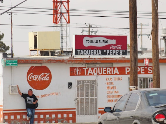 A street taco shop named 'Taqueria Pepe's' with a prominent Coca-Cola branding on its exterior. The building appears weathered and is painted primarily in white and orange. A person stands casually against the wall on the left, wearing jeans and a black t-shirt. There are several signs, including a green street sign reading 'Higuera'. Overhead, power lines are visible with a large red and white tower in the background. A gray car is parked in front on the right side.