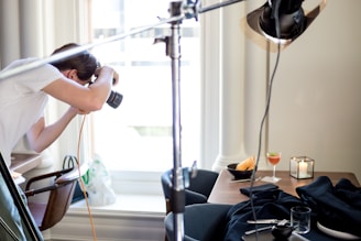 A person is leaning forward, focusing on taking a photo with a camera. The setup includes a table with a drink in a glass, a plate with food, and a lit candle, suggesting a food or product photography session. The scene is illuminated with professional lighting equipment, and there is a soft, natural light coming through a window.