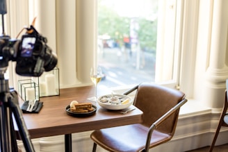 A dining setup on a wooden table includes a black plate with slices of brown bread, a bowl of soup with a spoon, and a glass of white wine. A camera on a tripod is positioned at the table, suggesting it is being photographed. The setting is well-lit by large windows, and decorative candles are also on the table.