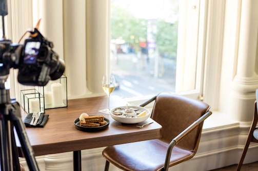 A dining setup on a wooden table includes a black plate with slices of brown bread, a bowl of soup with a spoon, and a glass of white wine. A camera on a tripod is positioned at the table, suggesting it is being photographed. The setting is well-lit by large windows, and decorative candles are also on the table.