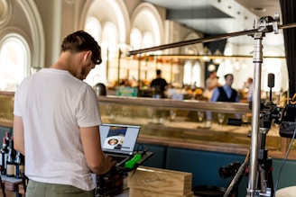 A man with short hair is working on a laptop in a stylish, contemporary restaurant setting. The background features arched windows and a bar area with bottles and glasses. The atmosphere is relaxed with warm lighting.