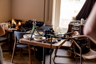 A dining table is set with several bowls of food, surrounded by chairs. A tripod with a camera is positioned on the table, suggesting a photo shoot of the meal. Nearby, other items like another camera, candles, and a handbag are visible. The setting has a cozy, modern interior with soft lighting.