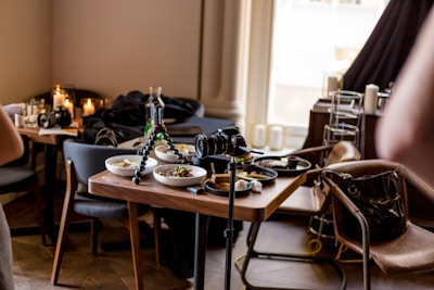 A dining table is set with several bowls of food, surrounded by chairs. A tripod with a camera is positioned on the table, suggesting a photo shoot of the meal. Nearby, other items like another camera, candles, and a handbag are visible. The setting has a cozy, modern interior with soft lighting.