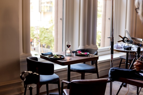 A small, elegantly set dining table for two is placed near large windows with tall, white curtains in an upscale setting. The table has plates of food, utensils, a lit candle, and a glass of red wine. There is a potted plant or tree visible outside the window. Additional chairs and a piece of equipment are visible in the background, suggesting this might be a behind-the-scenes photo or a part of a larger setup.