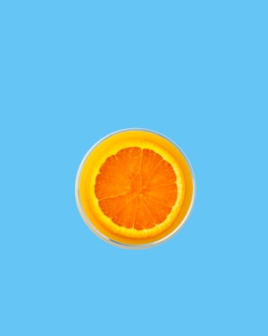 A vibrant orange slice is placed in the center of a clear glass filled with orange juice. The background is a solid light blue, creating a striking contrast with the orange hues.