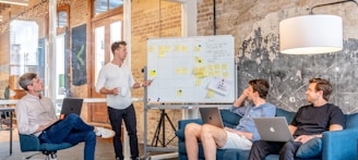 three men sitting while using laptops and watching man beside whiteboard