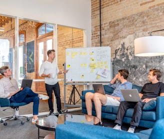 three men sitting while using laptops and watching man beside whiteboard