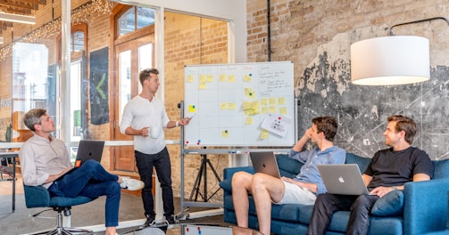 three men sitting while using laptops and watching man beside whiteboard