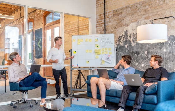 three men sitting while using laptops and watching man beside whiteboard