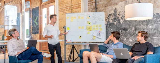 three men sitting while using laptops and watching man beside whiteboard