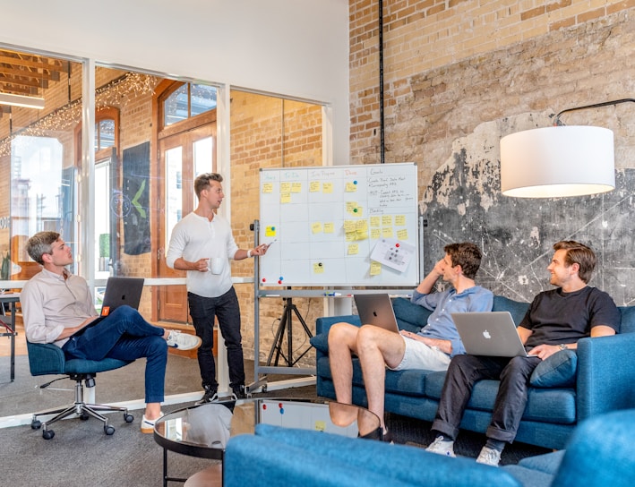 three men sitting while using laptops and watching man beside whiteboard