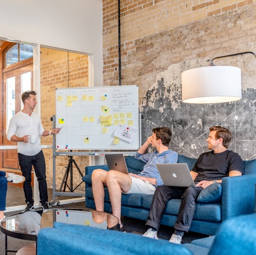 three men sitting while using laptops and watching man beside whiteboard