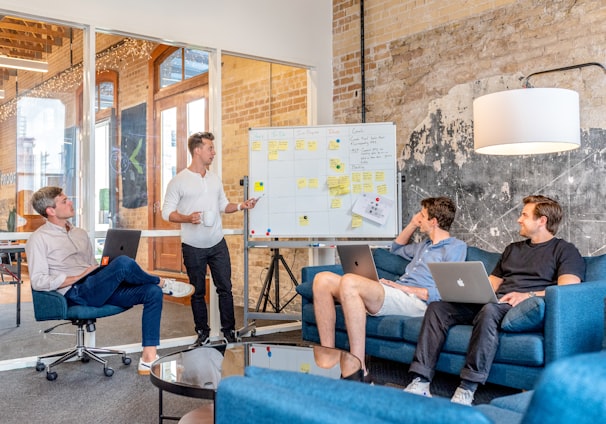 three men sitting while using laptops and watching man beside whiteboard