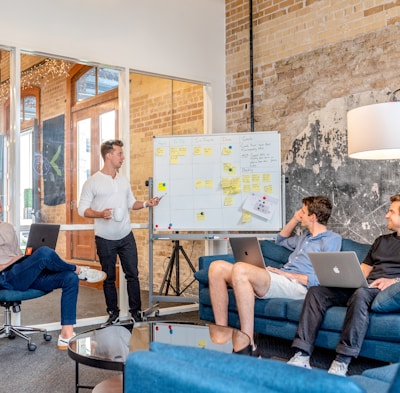 three men sitting while using laptops and watching man beside whiteboard