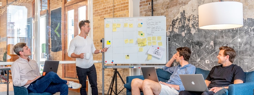 three men sitting while using laptops and watching man beside whiteboard