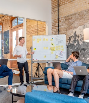 three men sitting while using laptops and watching man beside whiteboard