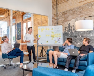 three men sitting while using laptops and watching man beside whiteboard