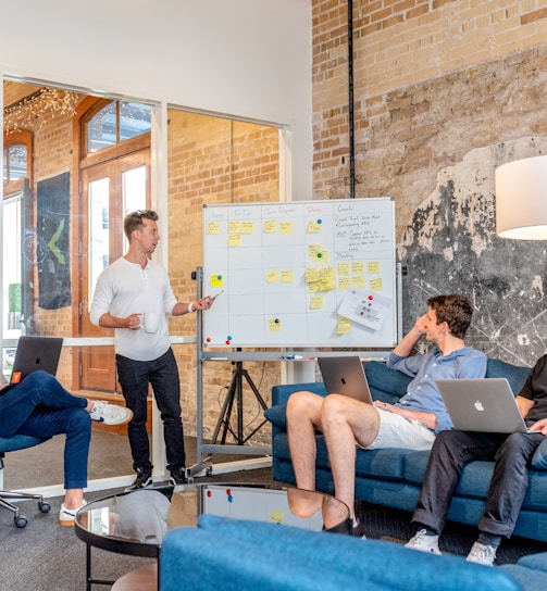 three men sitting while using laptops and watching man beside whiteboard