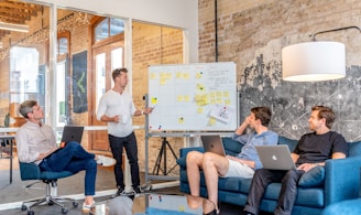 three men sitting while using laptops and watching man beside whiteboard