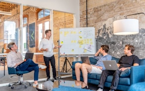 three men sitting while using laptops and watching man beside whiteboard