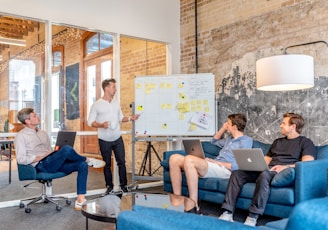 three men sitting while using laptops and watching man beside whiteboard