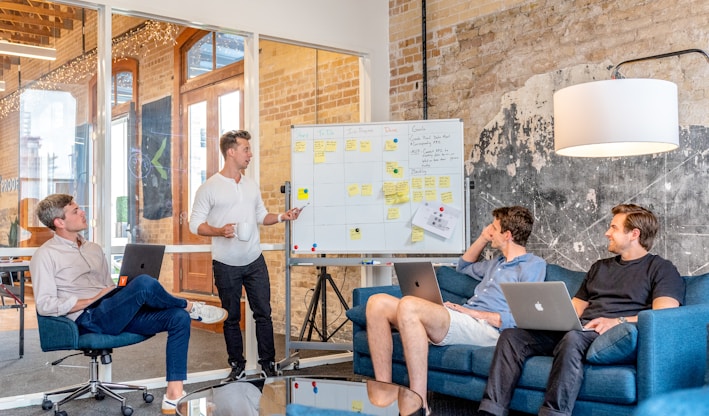 three men sitting while using laptops and watching man beside whiteboard