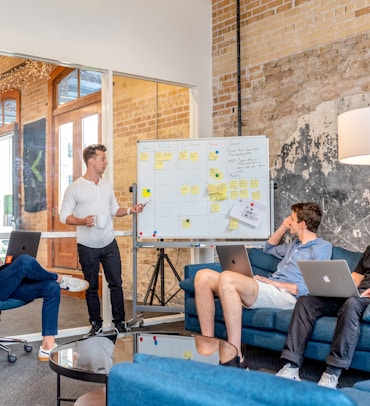 three men sitting while using laptops and watching man beside whiteboard