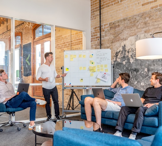 three men sitting while using laptops and watching man beside whiteboard