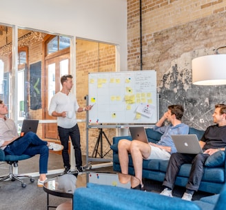 three men sitting while using laptops and watching man beside whiteboard