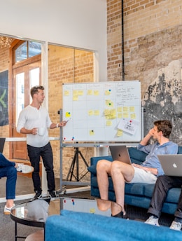 three men sitting while using laptops and watching man beside whiteboard