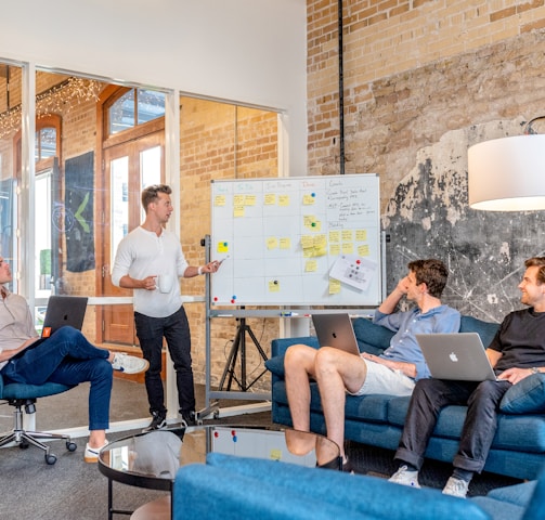 three men sitting while using laptops and watching man beside whiteboard