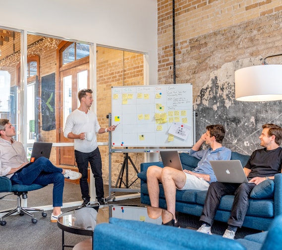 three men sitting while using laptops and watching man beside whiteboard