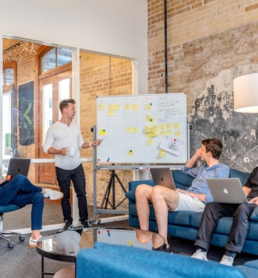 three men sitting while using laptops and watching man beside whiteboard