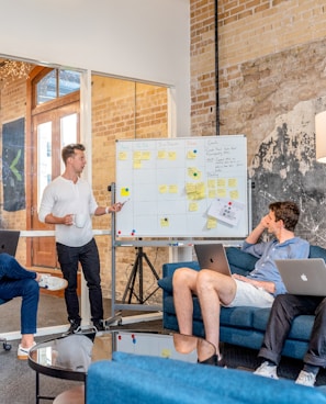 three men sitting while using laptops and watching man beside whiteboard