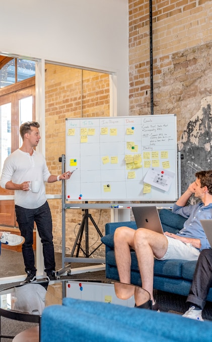 three men sitting while using laptops and watching man beside whiteboard