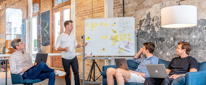 three men sitting while using laptops and watching man beside whiteboard