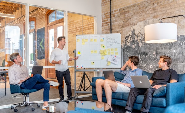 three men sitting while using laptops and watching man beside whiteboard