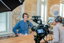 Two people are seated in a small recording studio. Both are wearing headphones and speaking into microphones, with one person facing a camera. The setup includes cameras, lights, and a computer monitor in the background. The room has brick walls and large windows allowing natural light in.
