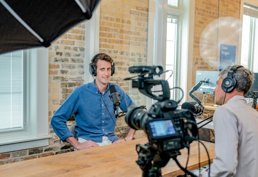 Two people are seated in a small recording studio. Both are wearing headphones and speaking into microphones, with one person facing a camera. The setup includes cameras, lights, and a computer monitor in the background. The room has brick walls and large windows allowing natural light in.