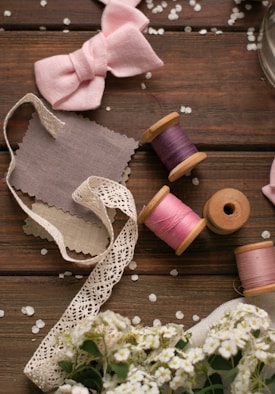 A flat lay of sewing and crafting materials includes pink and purple thread spools, delicate lace ribbon, fabric swatches, and a pink bow. The background is a wooden table scattered with white petals and small white flowers.
