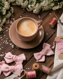 A warm cup of coffee sits on a wooden table surrounded by decorative pink fabric bows, lace, spools of thread, and scattered white flowers, creating a cozy, crafting atmosphere.